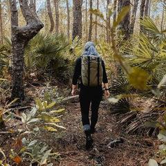 Wearing GORUCK’s Merino Challenge Socks, a hiker with a backpack treks through a lush forest surrounded by vibrant green foliage and tall trees.