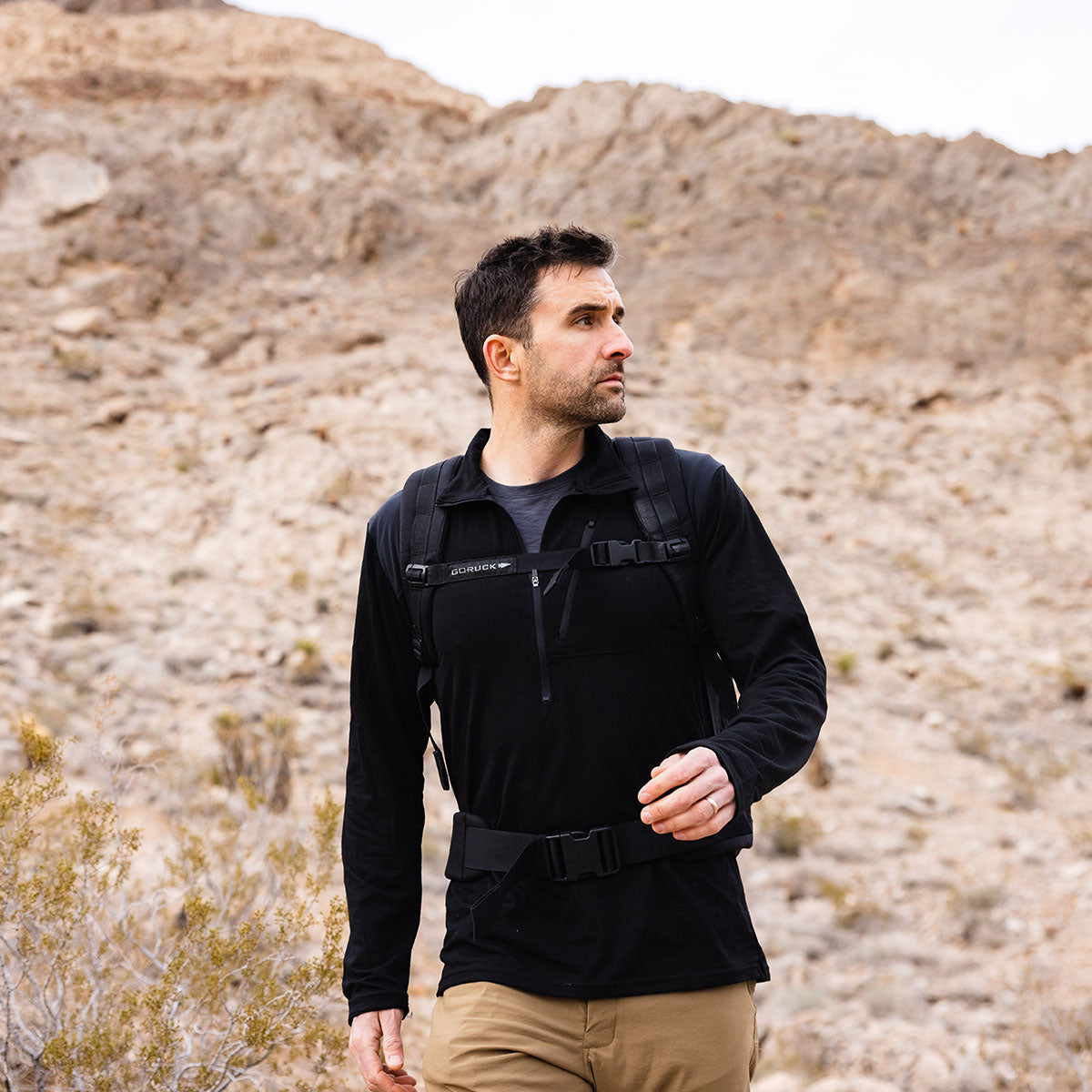 Man wearing a black jacket and backpack hiking in a rocky, desert landscape with mountains in the background.