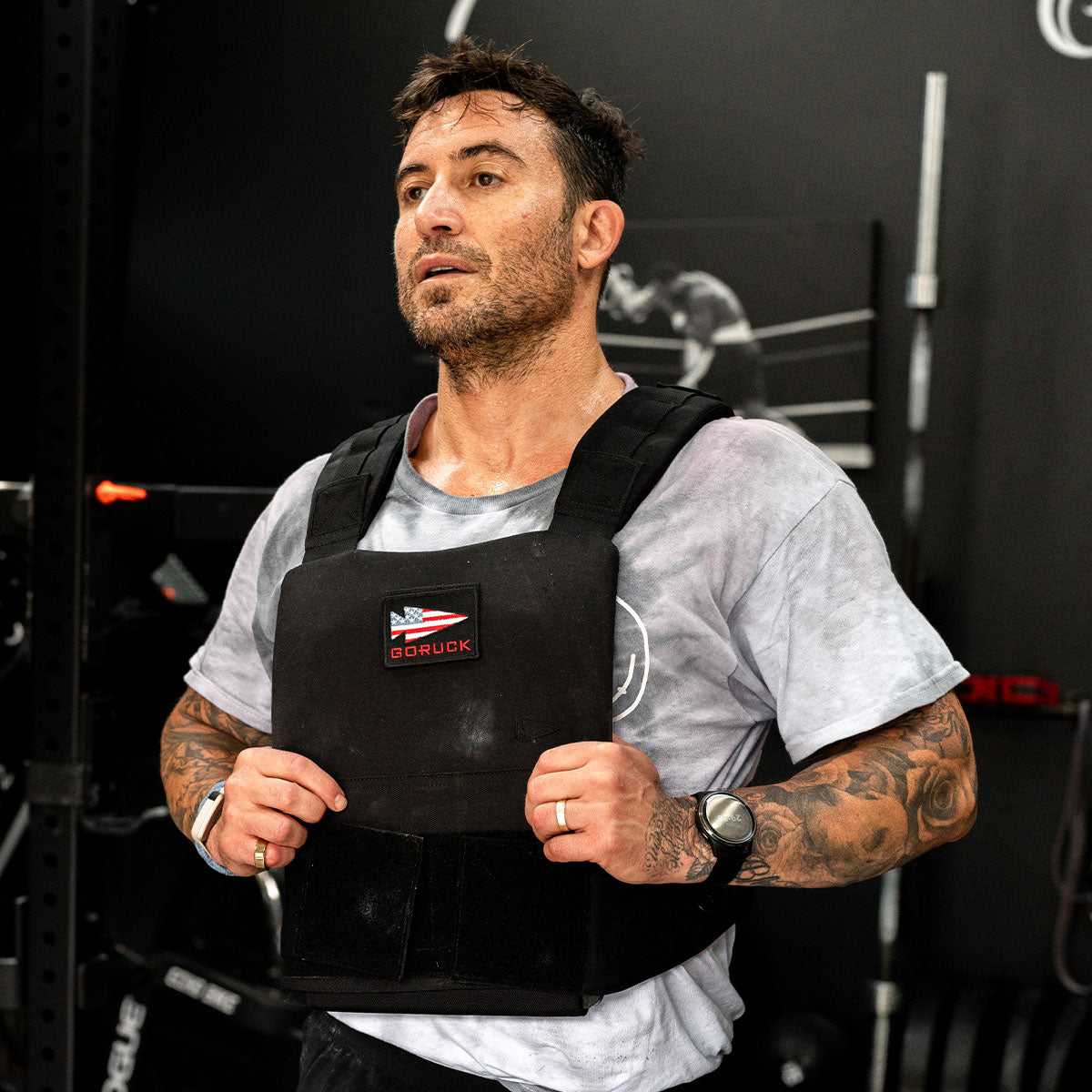Man with tattoos wearing a weighted vest and workout clothes, sweating during intense exercise indoors.