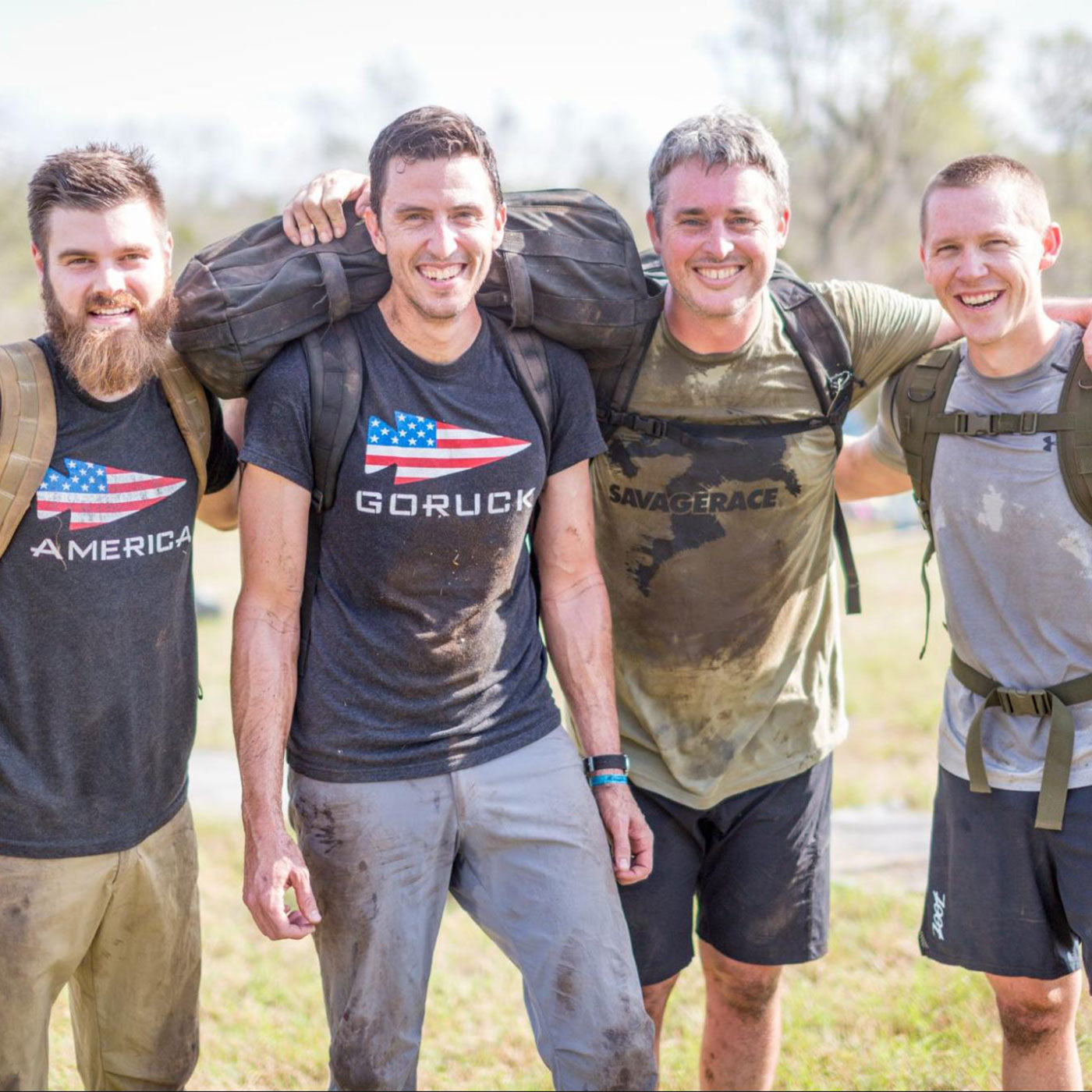 Four smiling men with muddy clothes and backpacks pose together outdoors after a workout or race.