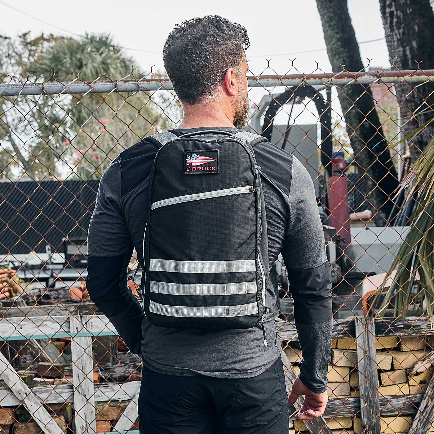 Man wearing a black and gray backpack stands outdoors near a chain-link fence and stacked firewood.