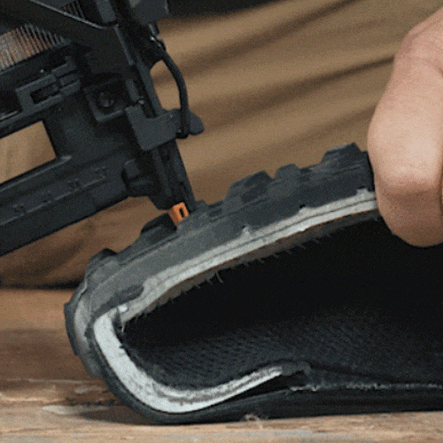 A hand uses a nail gun to attach a thick treaded shoe sole to black fabric on a wooden surface.