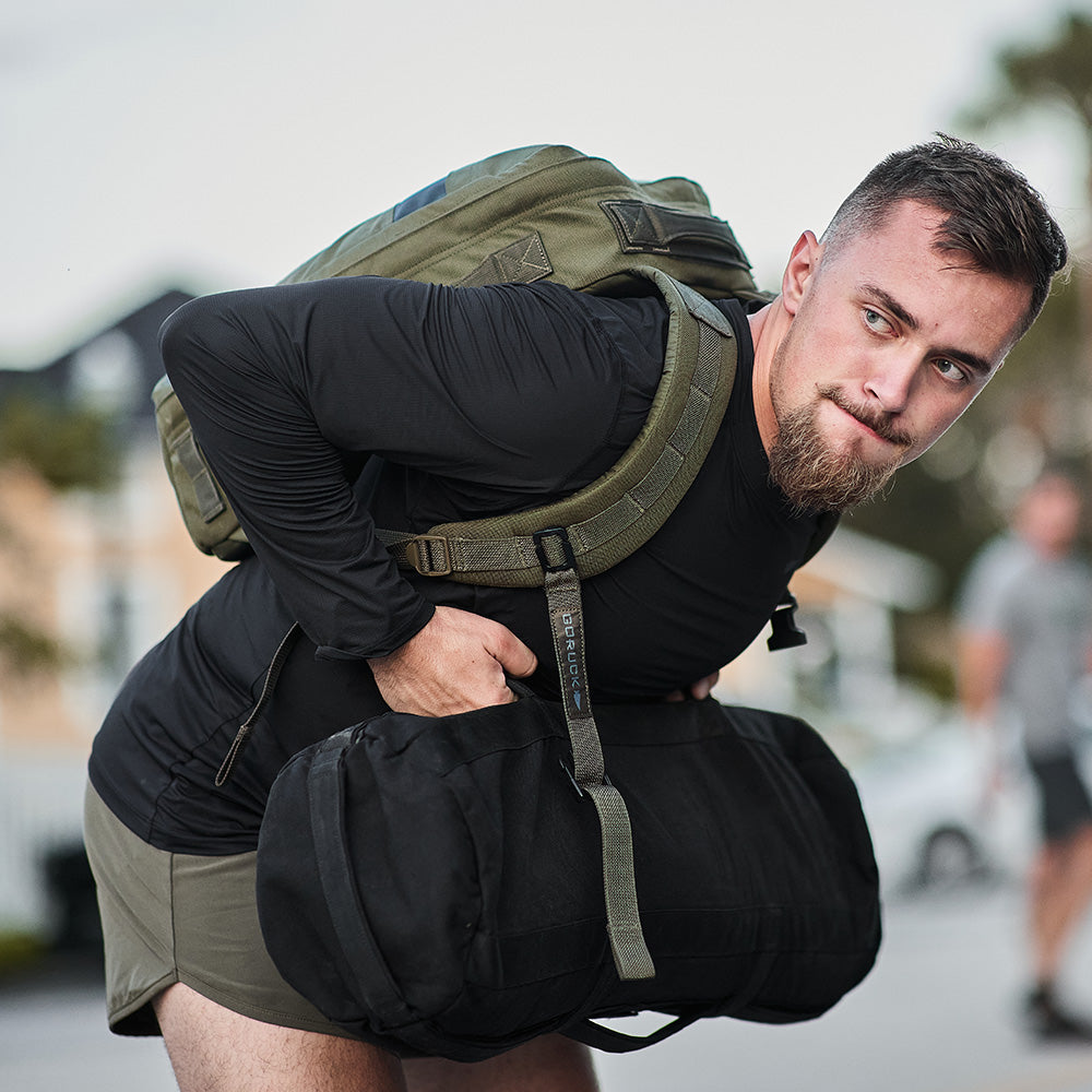 A man in athletic wear lifts a heavy duffel bag while carrying a backpack outdoors.