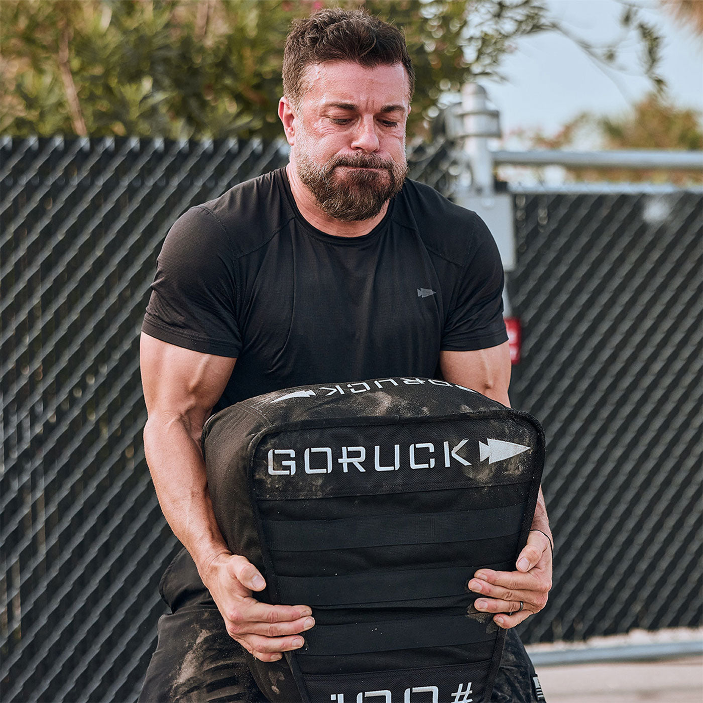 Man straining to lift a heavy GORUCK sandbag during an outdoor workout near a chain-link fence.