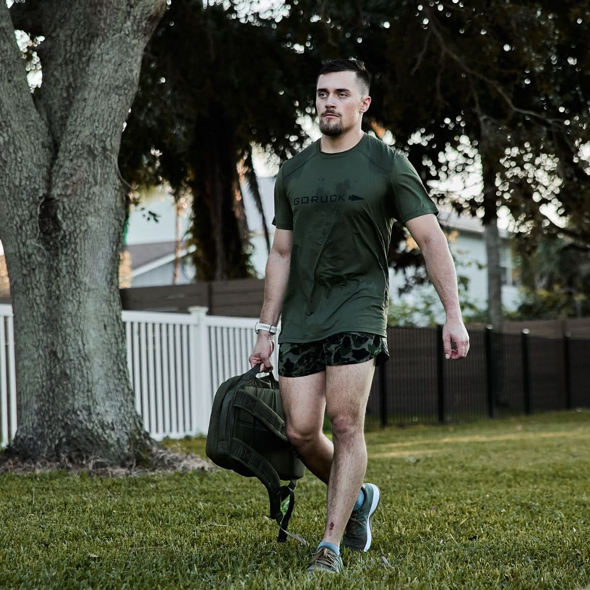 Man wearing olive GORUCK Tough Mesh Tee, carrying rucksack outdoors on grass