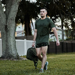 Man wearing olive GORUCK Tough Mesh Tee, carrying rucksack outdoors on grass