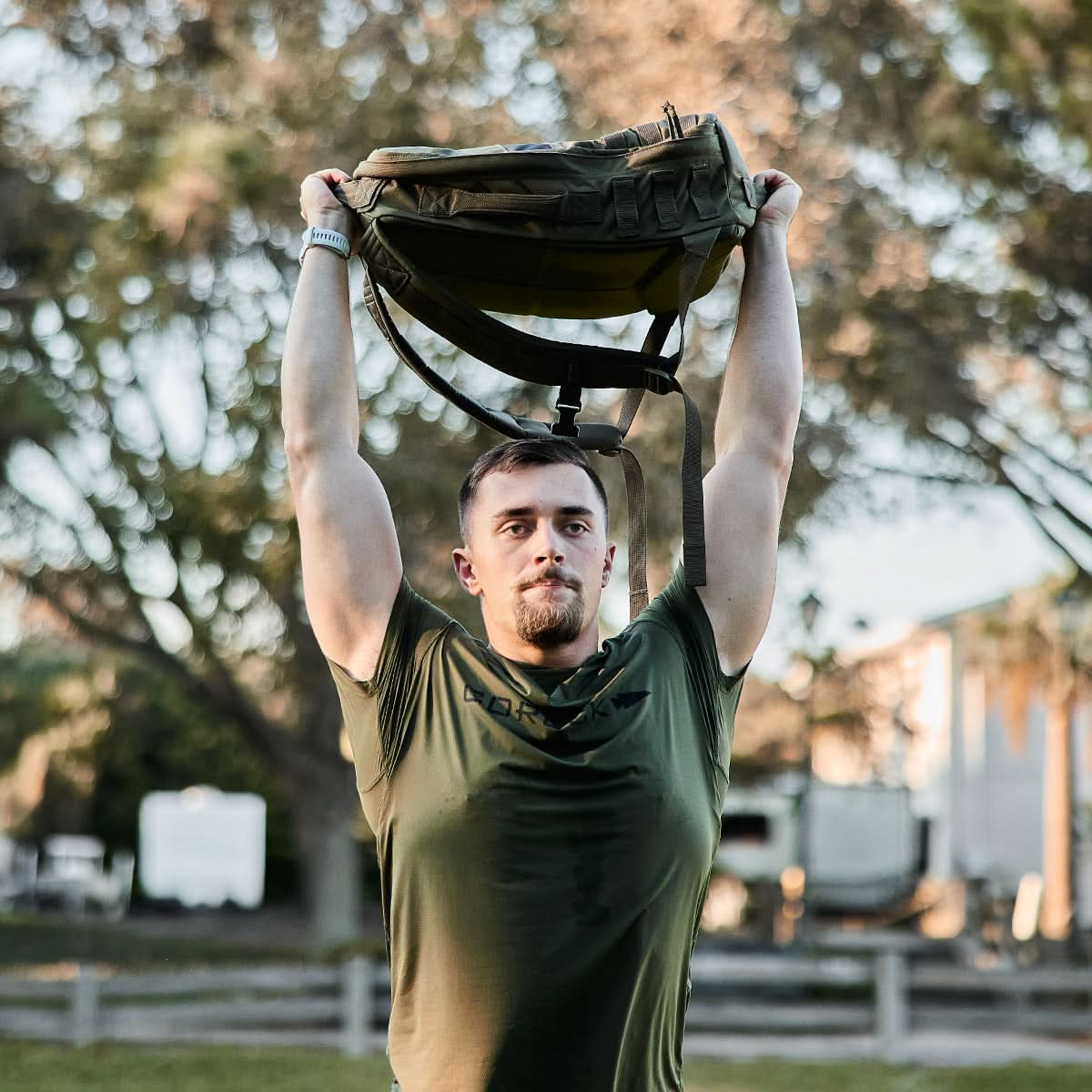 Man outdoors lifting rucksack overhead wearing olive GORUCK Tough Mesh Tee