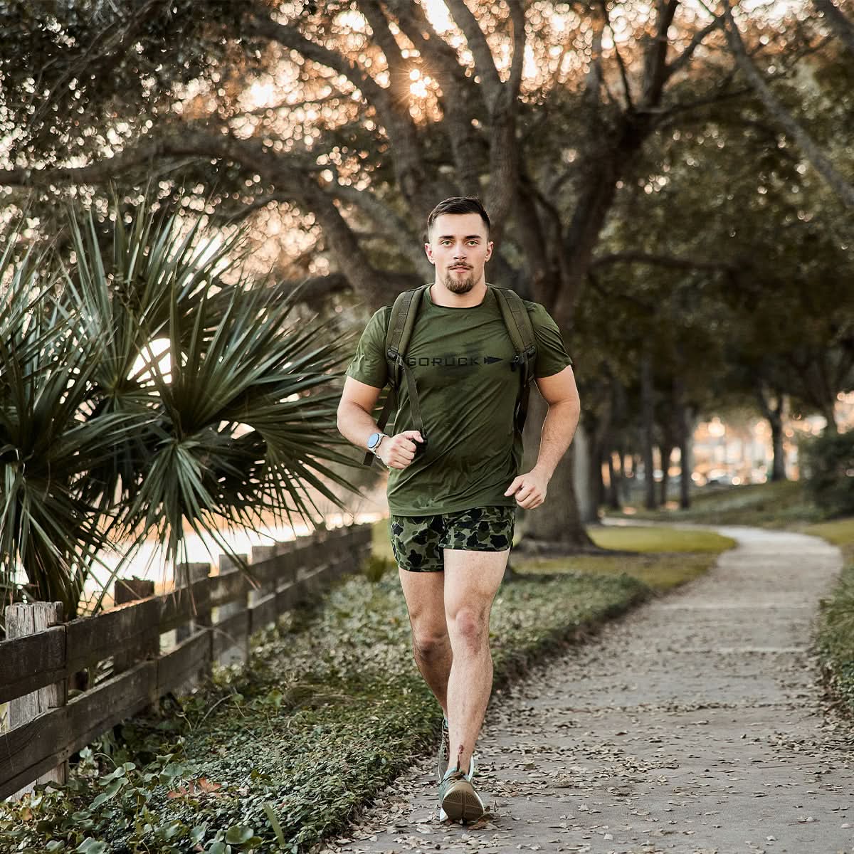 Man rucking in GORUCK olive Tough Mesh Tee outdoors, wearing rucksack on tree-lined path