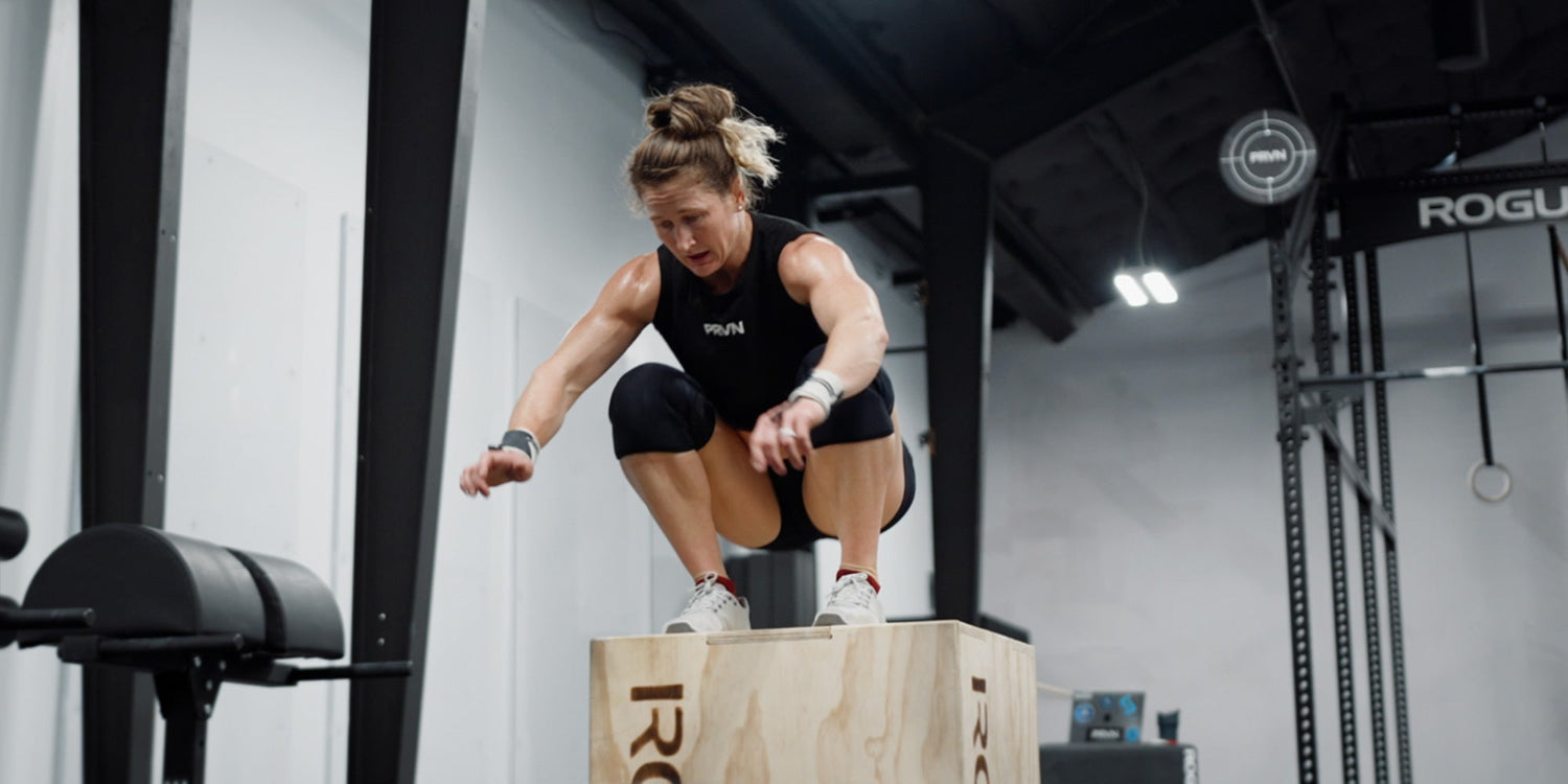 Woman in athletic wear doing a box jump exercise in a gym, landing on a wooden box.