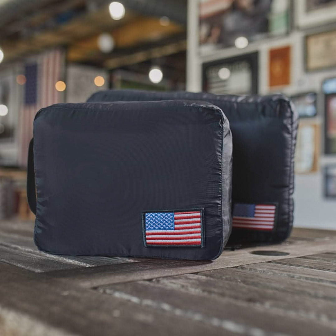 Two black GORUCK rucks with American flag patches, crafted from tough 200D nylon and ideal for use with Packing Cubes, sit on a wooden table in a room decorated with framed art.