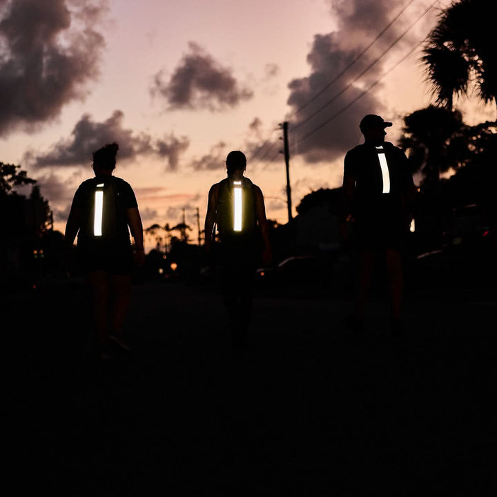 Three people with reflective strips on their Basic Rucker® backpacks walk at dusk beneath a cloudy sky and silhouetted palm trees.