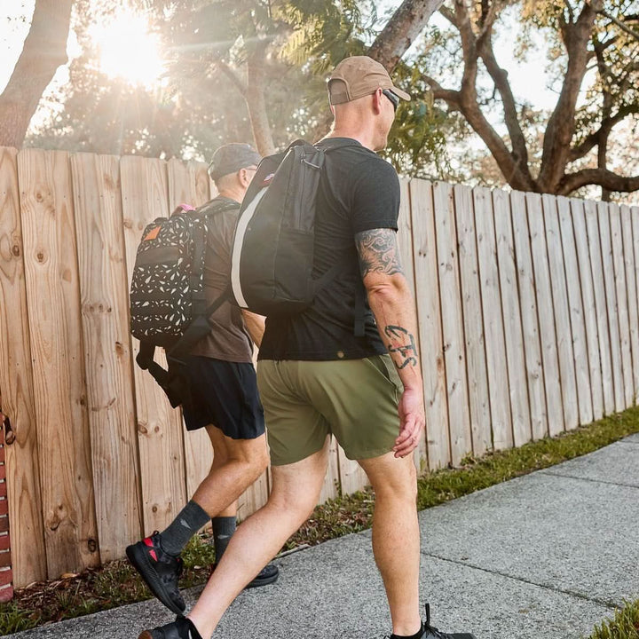 Two men rucking outdoors with GORUCK backpacks, walking on a sidewalk by a wooden fence