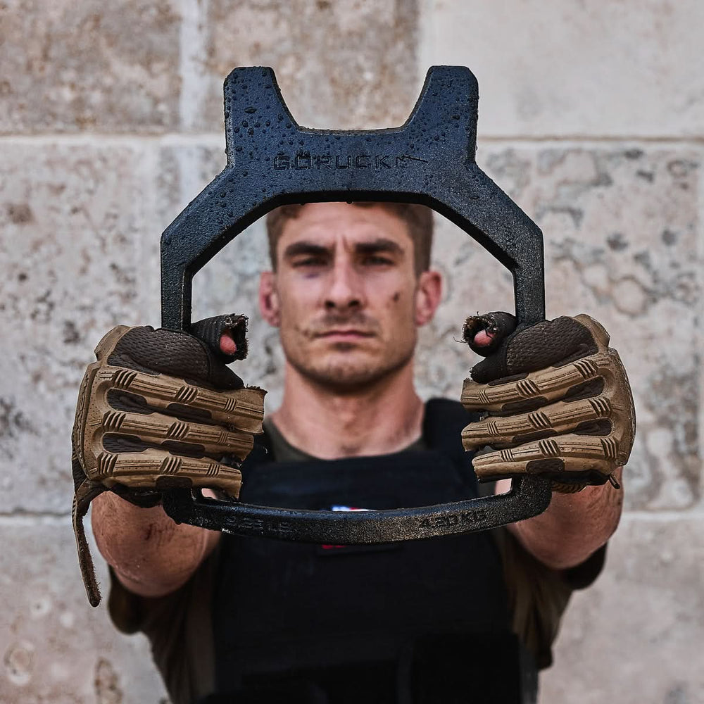 A man wearing tactical gloves holds a Curved Ruck Plate, labeled "GORUCK," while standing against a stone wall.