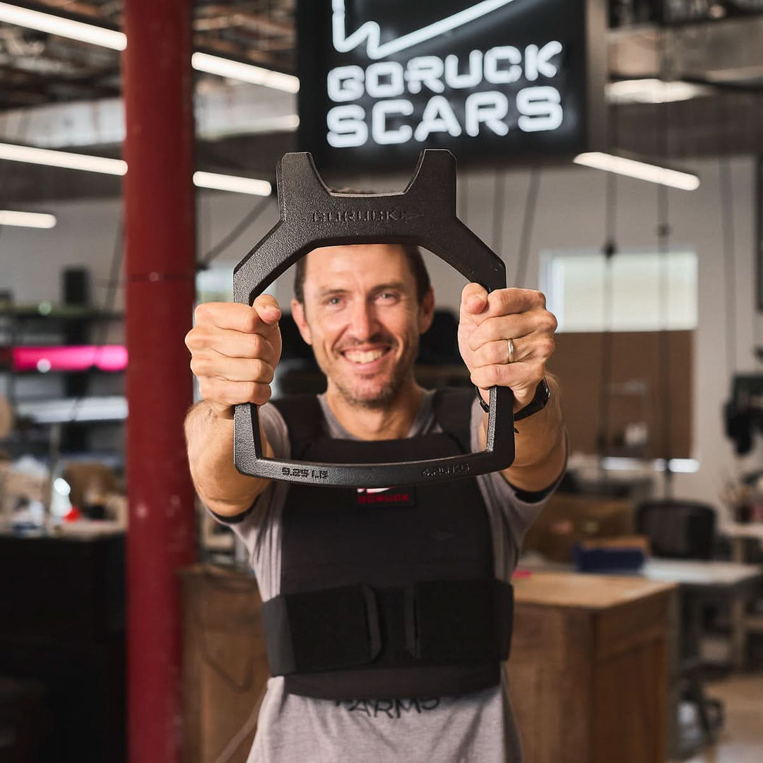 Smiling man in a workshop holds up a black Rucking Weight Vest, with a "GORUCK SCARS" sign visible in the background.