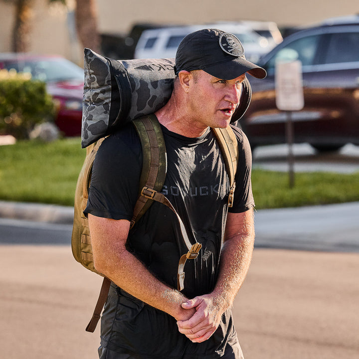 A man in a black shirt and cap, carrying a backpack, camouflage mat, and GORUCK Simple Training Sandbags, walks outdoors on a sunny day.