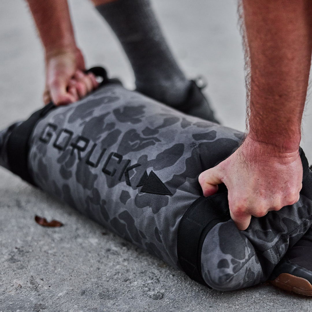 A person grips a GORUCK Simple Training Sandbag in camouflage on concrete, preparing to lift it during a home gym workout.
