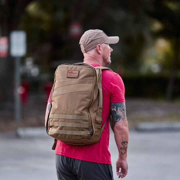A man in a red shirt and tan cap wears the GORUCK Rucker 4.0 outdoors, facing away from the camera.
