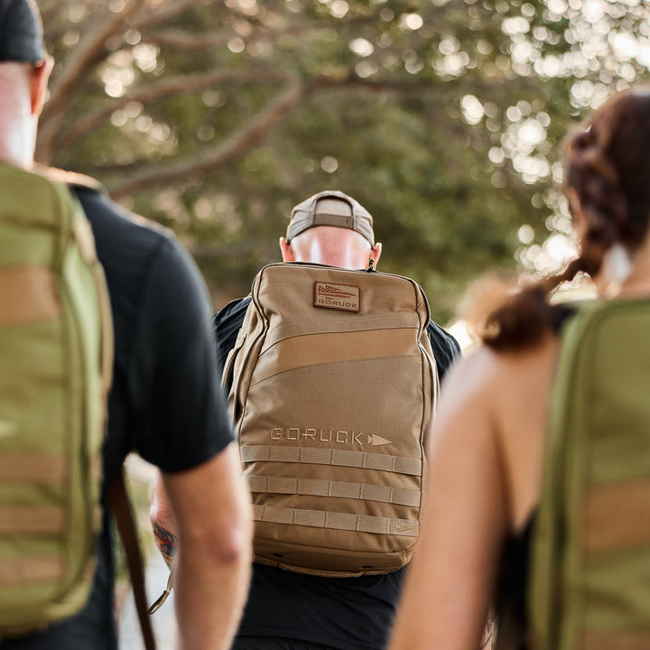 Three people outdoors wear backpacks, with a focus on the tan GORUCK Rucker 4.0 bag in the center.