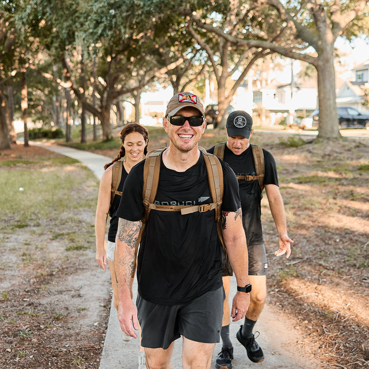 Three people carrying GORUCK Rucker 4.0 backpacks walk and smile along a shaded outdoor path lined with trees and houses.