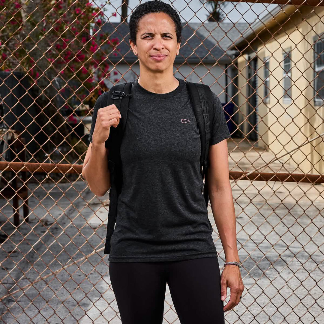 A person wearing the vendor-unknown Subtle Spearhead Tee - Tri-Blend in black and a backpack stands outdoors by a chain-link fence, with the GORUCK spearhead patch visible.