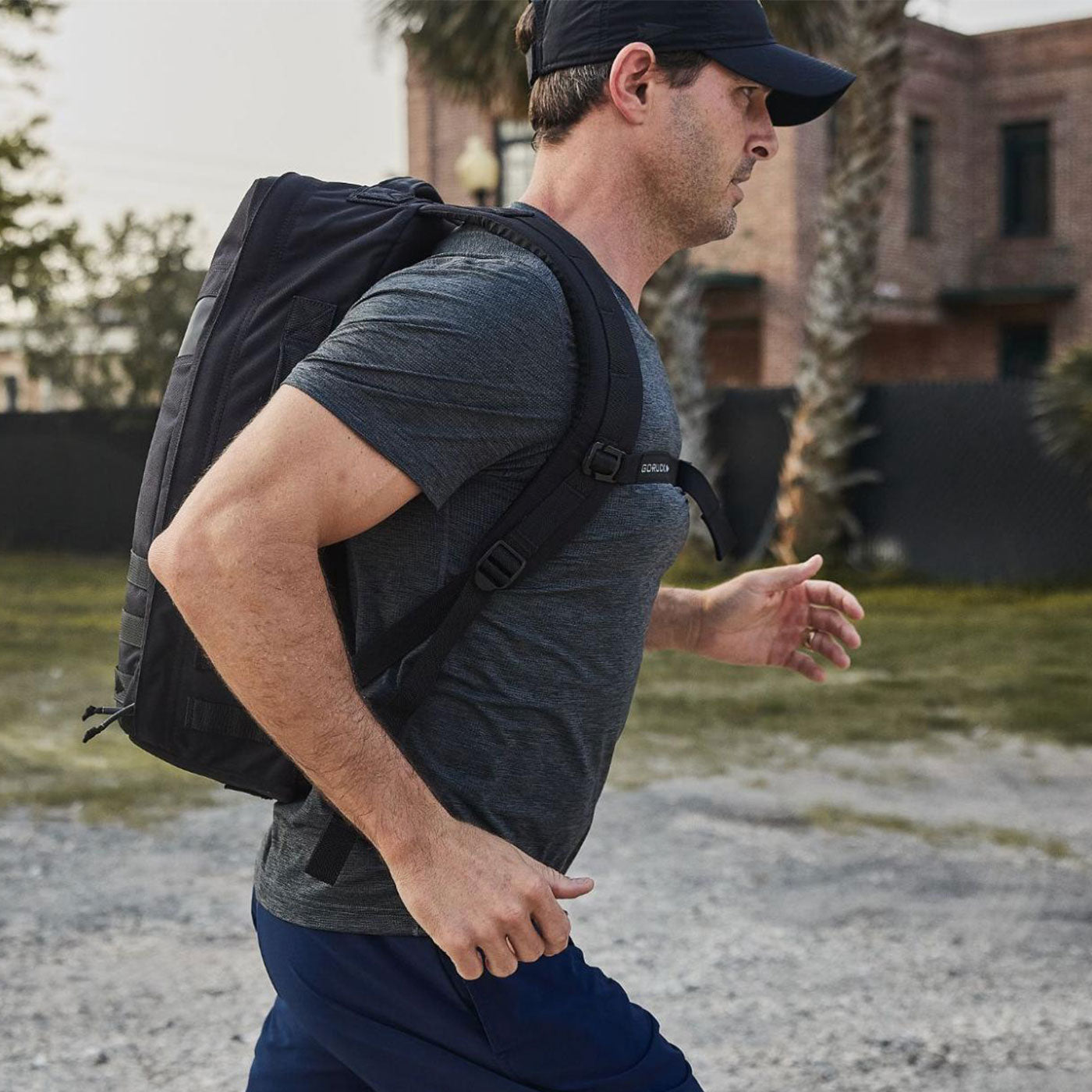 Man in athletic clothing and a cap jogging outdoors with a black backpack in an urban area.