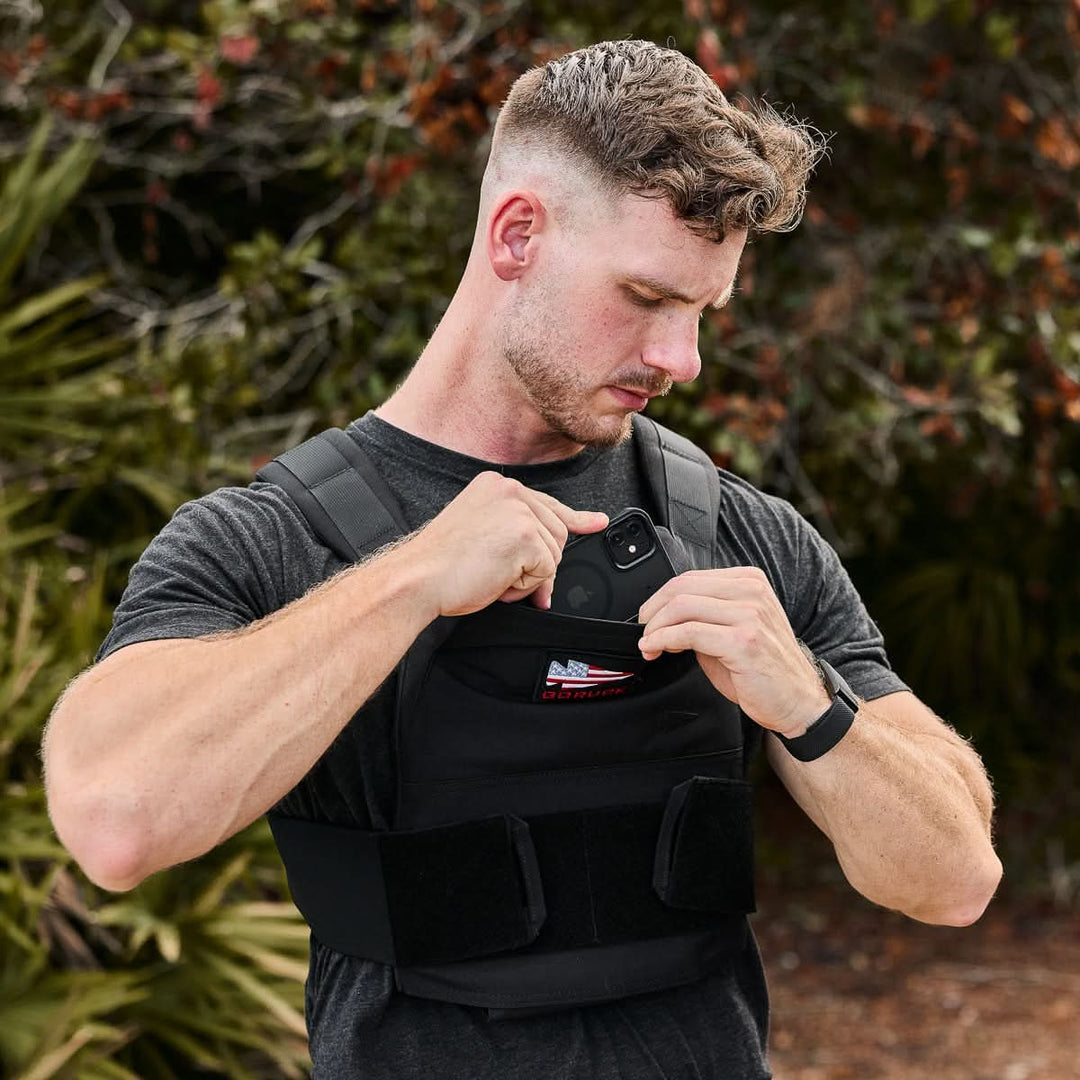 A man outdoors with greenery in the background puts a smartphone into the front pocket of a black Rucking Weight Vest.