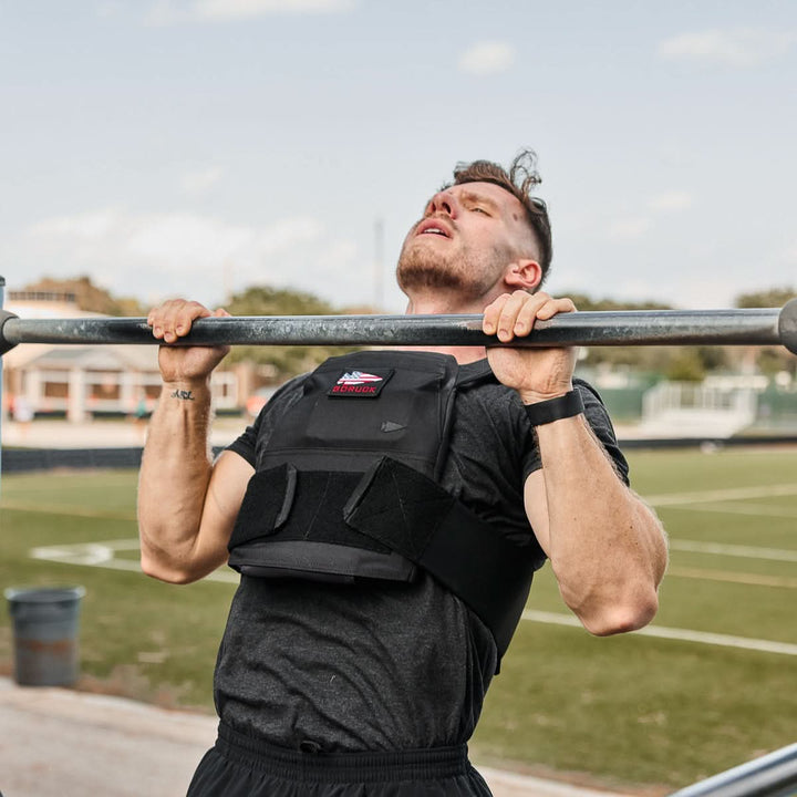 A man wearing the Rucking Weight Vest with Curved Ruck Plates does a pull-up on an outdoor bar, with a field visible in the background.