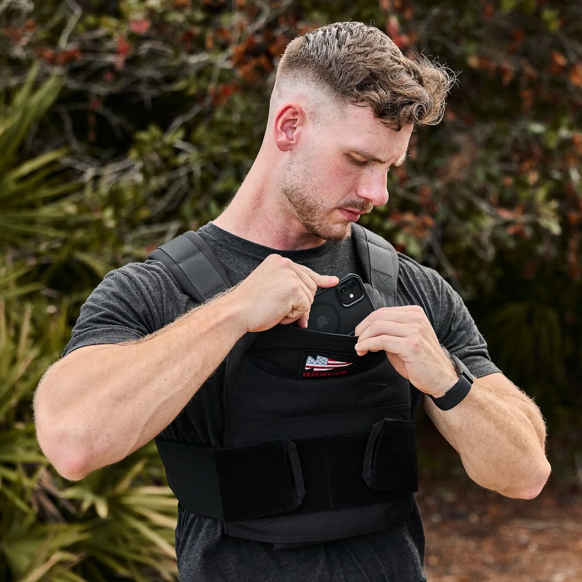 Man wearing GORUCK weighted training vest outdoors, placing smartphone in chest pocket