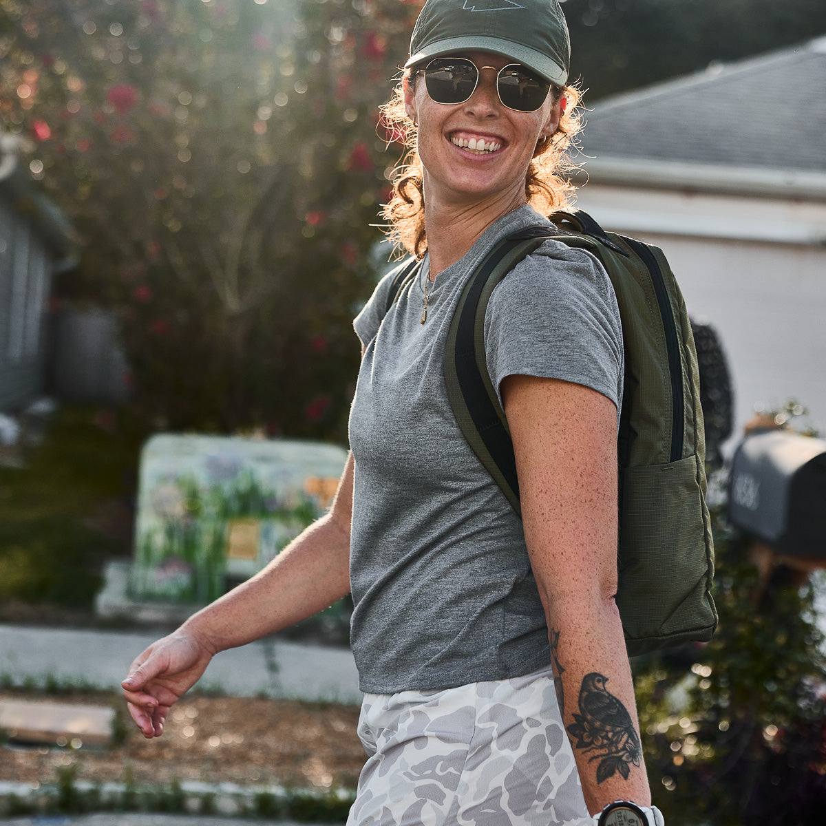 Smiling person in a gray t-shirt and cap walks outdoors with a backpack, sunlight in the background.