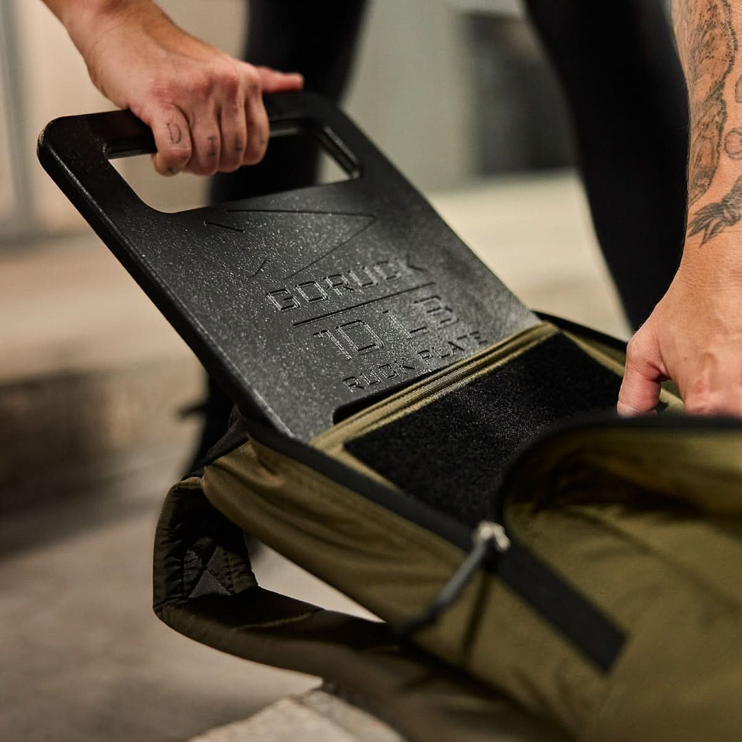 A person puts a 15 lb "Born Primitive" workout plate into an olive green Basic Rucker®, turning it into a weighted rucksack for training.
