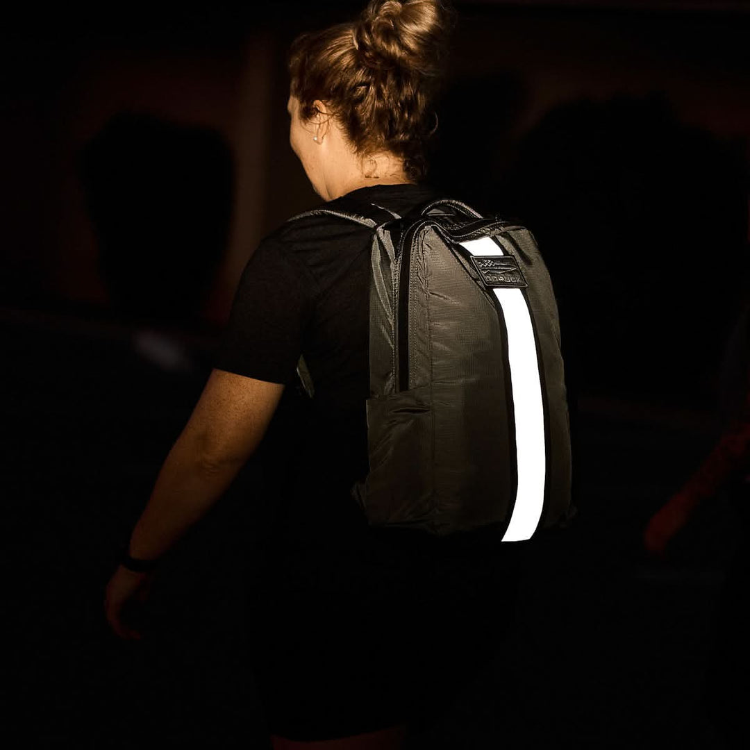 A person with a bun hairstyle wears a dark Basic Rucker® backpack with a reflective strip down the center.