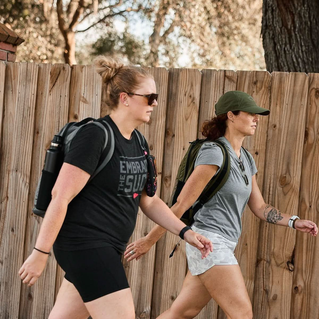 Two women rucking with GORUCK backpacks outdoors by a wooden fence