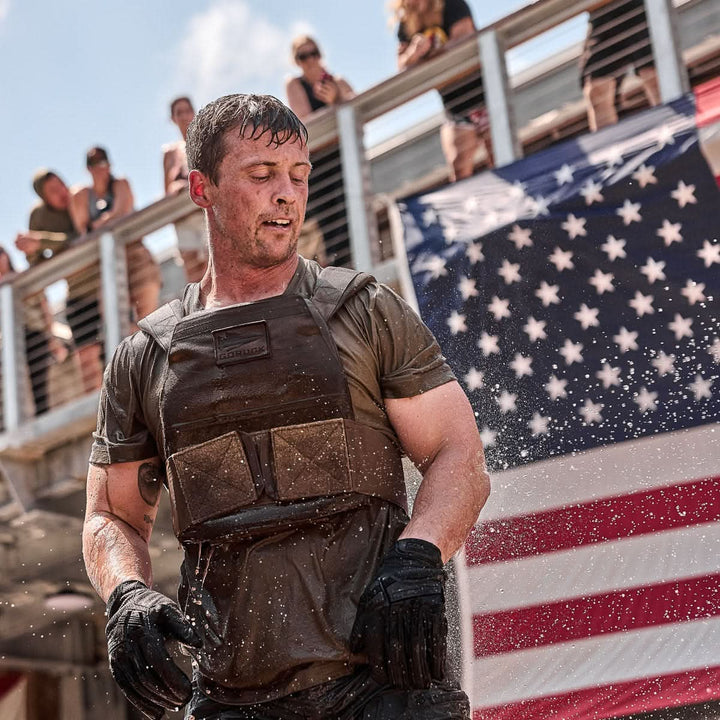 A man wearing the Rucking Weight Vest and wet tactical gear stands before a large American flag, as onlookers watch from above.