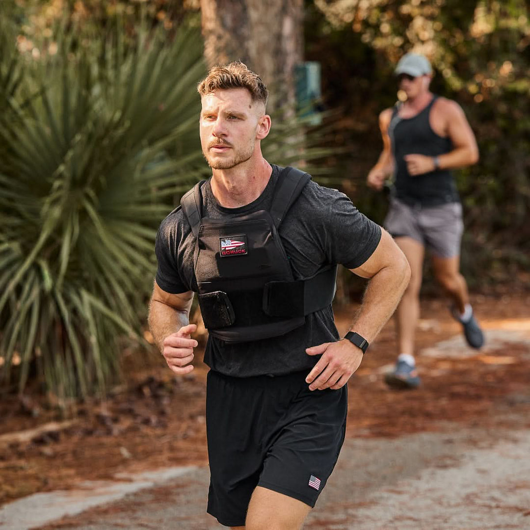 A man runs on a wooded trail wearing a Rucking Weight Vest with Curved Ruck Plates, while another runner follows behind, both embracing the intensity of rucking.