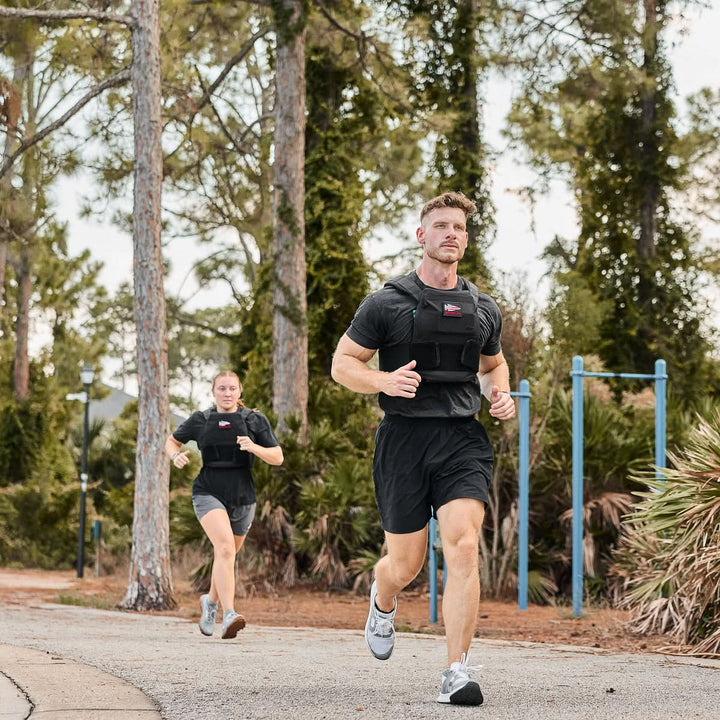 Two people jog outdoors on a tree-lined path, both dressed in black workout gear and wearing weight vests loaded with Curved Ruck Plates for added resistance.