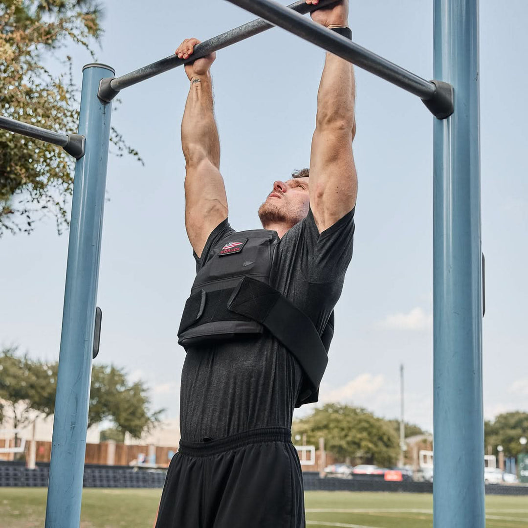 A man wearing a weight vest with Curved Ruck Plates does a pull-up on outdoor bars in a park on a sunny day.