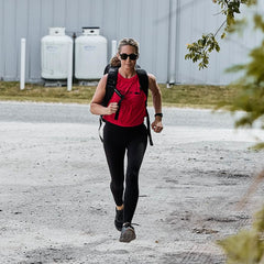 Wearing a Women’s Performance Tank - ToughMesh from GORUCK, black leggings, and sunglasses, a person jogs briskly along a gravel path with a backpack. With greenery and propane tanks in the background, they move with precision as if training for a trek through the Italian Alps.