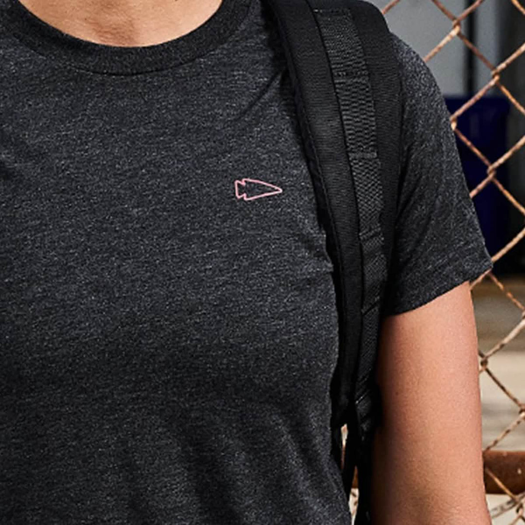 Wearing the Subtle Spearhead Tee - Tri-Blend by vendor-unknown in dark gray, featuring a subtle spearhead logo, and carrying a black backpack while standing by a chain-link fence.