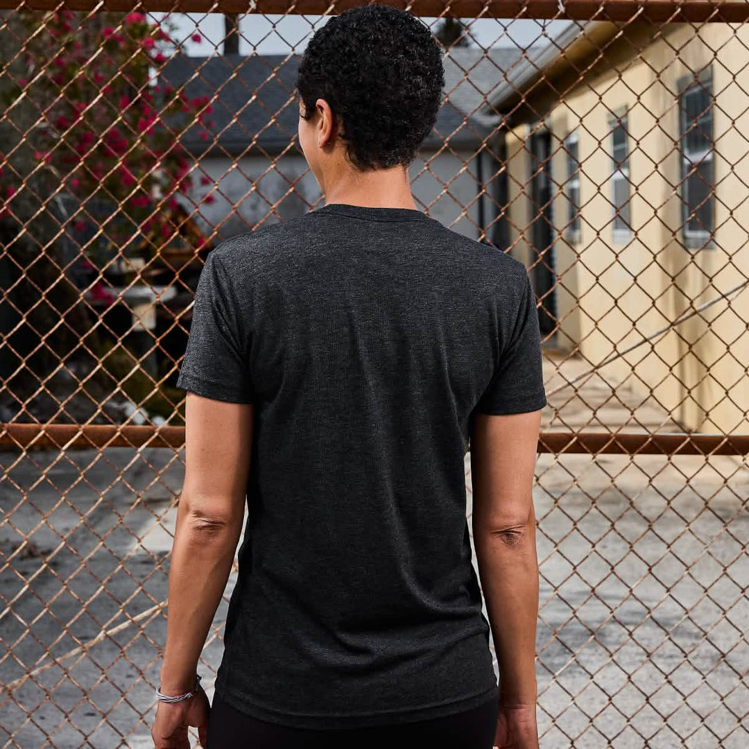 A person with short hair wears the Subtle Spearhead Tee - Tri-Blend by vendor-unknown in dark color, standing outdoors by a chain-link fence.