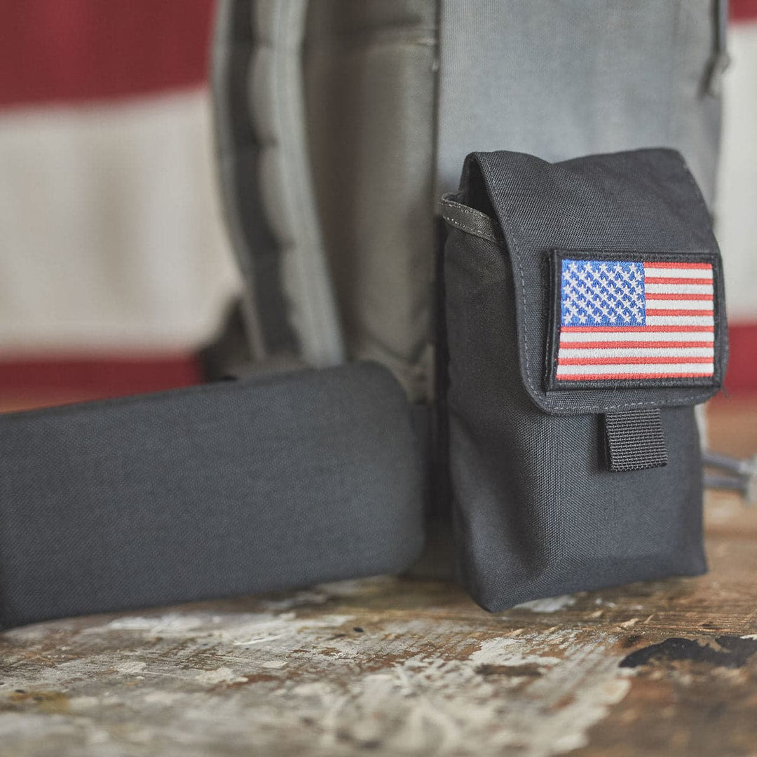 A GORUCK black pouch adorned with a US flag patch rests on a wooden surface beside a rugged GORUCK backpack with a Padded Hip Belt, set against a blurred flag background. Crafted from durable CORDURA fabric, it offers resilience and style for adventurous rucks.