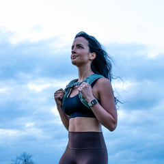 A woman in workout clothes wears the Spy Ruck | Women's Weighted Vest outdoors, looking upward against a cloudy sky.