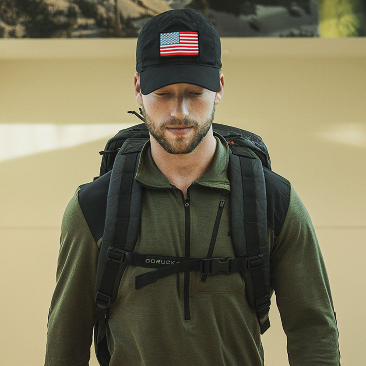 Man wearing a black cap with an American flag and a large black backpack, looking down.