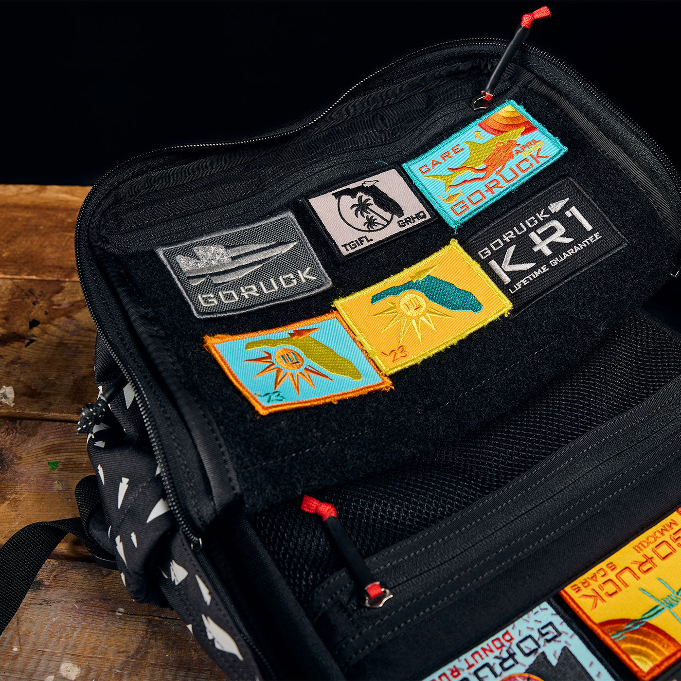 Open backpack displaying colorful embroidered patches on a velcro panel, resting on a wooden surface.
