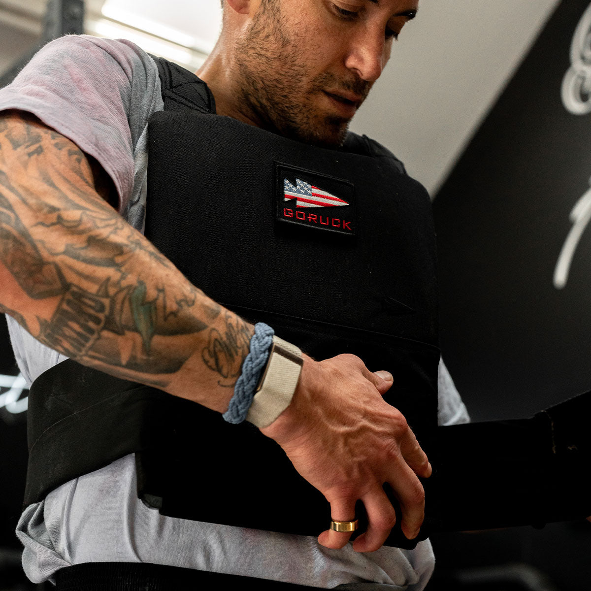 A tattooed man puts on a black GORUCK weighted vest in a gym setting.