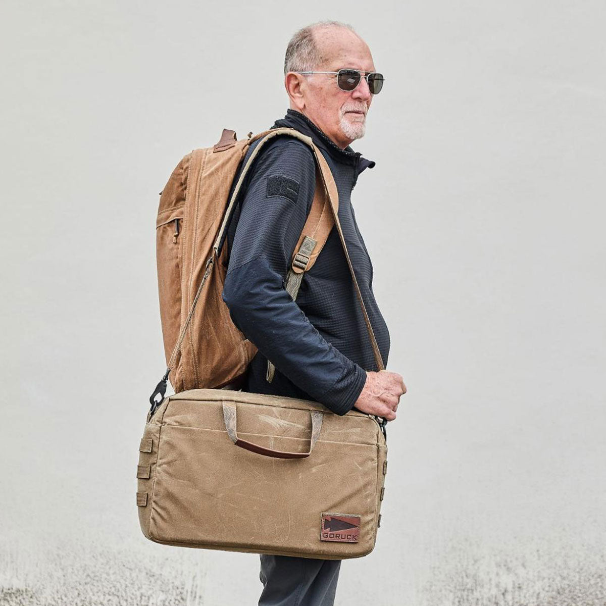 Older man with sunglasses carrying a tan backpack and matching shoulder bag, standing against a plain background.