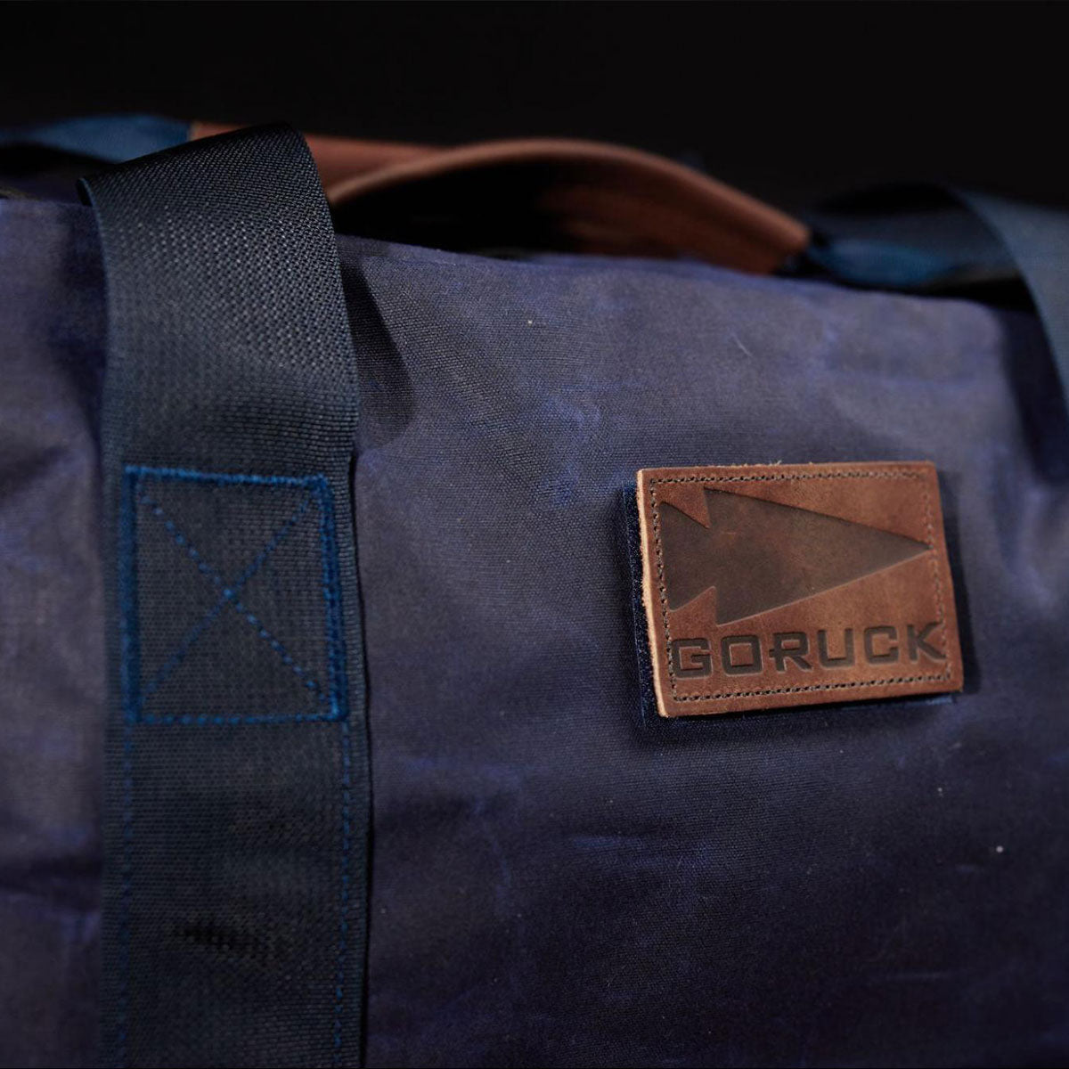 Close-up of a dark blue bag with black handles and a brown leather patch labeled “GORUCK.”.