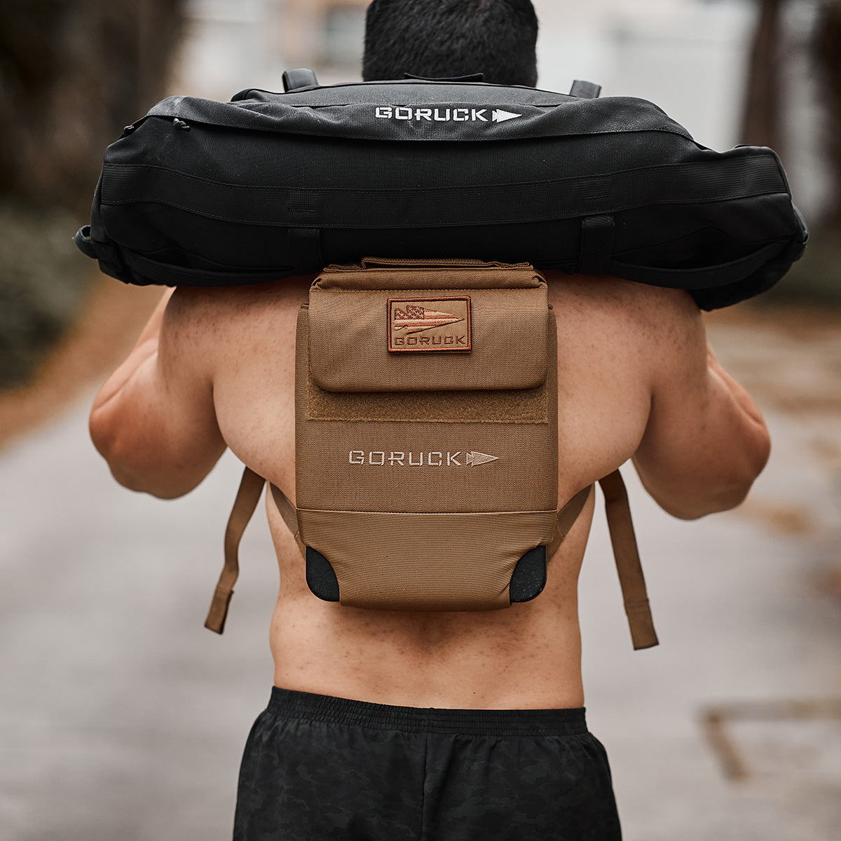 A person carries a black Ruck Plate Carrier 3.0 sandbag while their tan backpack is crafted from durable ballistic nylon.