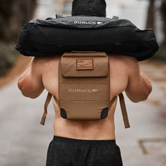 A person carries a black Ruck Plate Carrier 3.0 sandbag while their tan backpack is crafted from durable ballistic nylon.