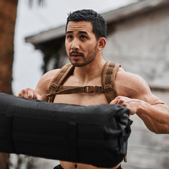 A person with a Ruck Plate Carrier 3.0, featuring ergonomic lumbar support, lifts a heavy bag outdoors, looking focused.