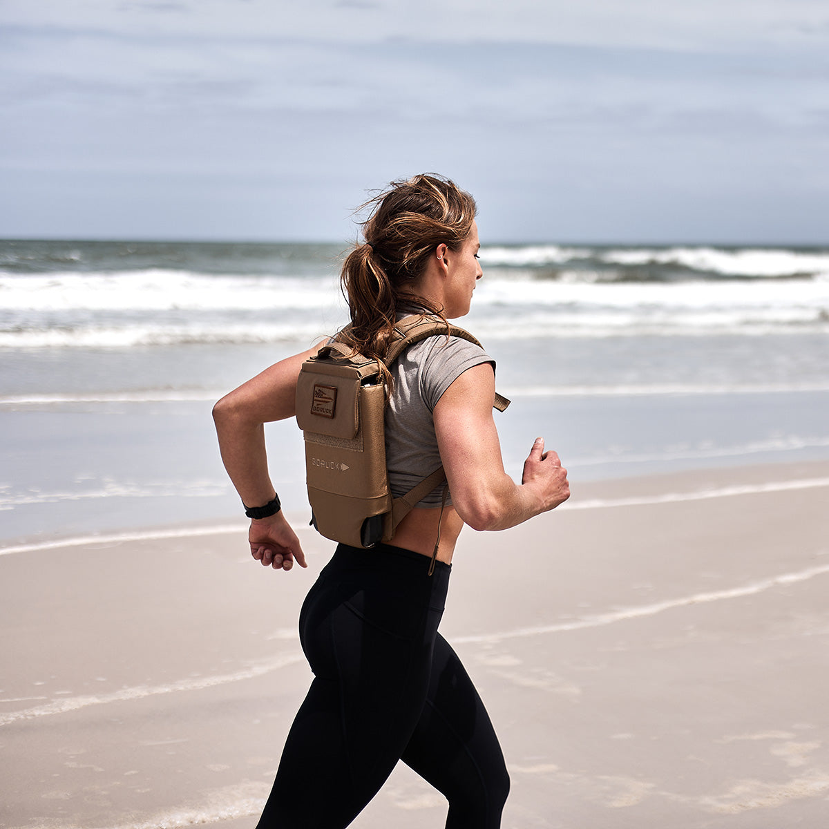 A woman runs on a sandy beach with the Ruck Plate Carrier 3.0 backpack, featuring ergonomic lumbar support, as waves crash in the background.